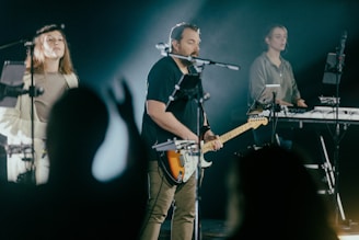 A group of musicians performing on stage, featuring a guitarist in the center, a female vocalist on one side, and a keyboard player on the other. They are surrounded by various music equipment and microphones. The lighting is dim with a spotlight on the performers, creating a focused and dynamic atmosphere.
