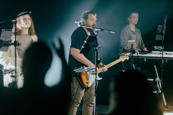 A group of musicians performing on stage, featuring a guitarist in the center, a female vocalist on one side, and a keyboard player on the other. They are surrounded by various music equipment and microphones. The lighting is dim with a spotlight on the performers, creating a focused and dynamic atmosphere.