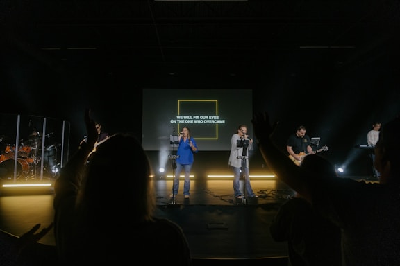 A band is performing on stage with a screen displaying the text 'WE WILL FIX OUR EYES ON THE ONE WHO OVERCAME.' Several musicians, including guitarists and a keyboard player, are visible under stage lighting. The audience can be seen silhouetted in the foreground, with some members raising their hands.