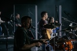 A music band is performing on stage, featuring musicians playing drums, guitar, and keyboard. They are surrounded by transparent screens and microphones, with focused expressions and illuminated by stage lights.
