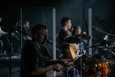 A music band is performing on stage, featuring musicians playing drums, guitar, and keyboard. They are surrounded by transparent screens and microphones, with focused expressions and illuminated by stage lights.