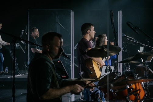 A music band is performing on stage, featuring musicians playing drums, guitar, and keyboard. They are surrounded by transparent screens and microphones, with focused expressions and illuminated by stage lights.