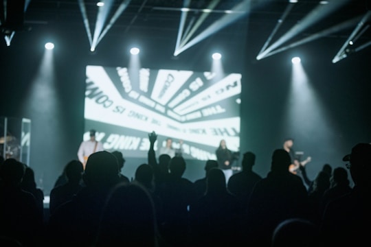 A dimly lit concert or event setting featuring a stage with performers and illuminated by spotlights. The audience is mostly in silhouette in the foreground, with a large screen in the background displaying abstract graphics.