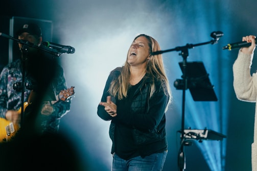 A woman clapping and singing energetically on stage, surrounded by musicians and microphones. The stage is illuminated with blue lighting, creating a vibrant atmosphere.