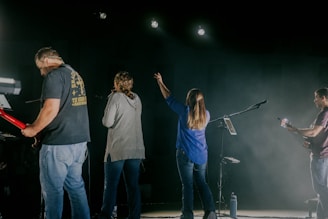 Four musicians stand on a dimly lit stage, each engaged with their instruments. One plays a red electric guitar, while another has a microphone. A woman raises her hand towards the bright stage lights above, and another person is captured in mid-performance. Microphones and stands are visible, highlighting a live music setup.