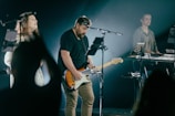 Musicians are performing on stage with a focus on a guitarist playing an electric guitar. A female vocalist and another individual playing a keyboard are visible. The stage is dimly lit, creating a spotlight effect on the performers.