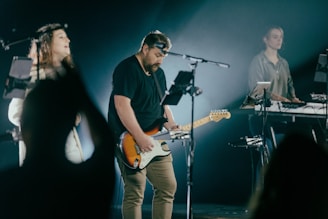 Musicians are performing on stage with a focus on a guitarist playing an electric guitar. A female vocalist and another individual playing a keyboard are visible. The stage is dimly lit, creating a spotlight effect on the performers.