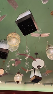 A decorative ceiling features various hanging light fixtures and ornamental designs. The lights include vintage-style glass and metal lanterns, surrounded by artistic representations of insects and birds. The ceiling is painted green, adding a unique backdrop to the eclectic arrangement.