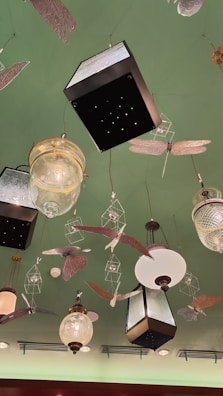 A decorative ceiling features various hanging light fixtures and ornamental designs. The lights include vintage-style glass and metal lanterns, surrounded by artistic representations of insects and birds. The ceiling is painted green, adding a unique backdrop to the eclectic arrangement.