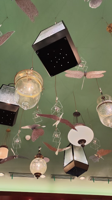 A decorative ceiling features various hanging light fixtures and ornamental designs. The lights include vintage-style glass and metal lanterns, surrounded by artistic representations of insects and birds. The ceiling is painted green, adding a unique backdrop to the eclectic arrangement.