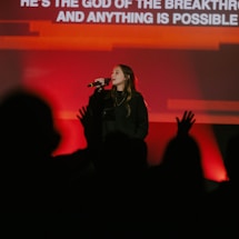 A female performer is singing into a microphone on stage. Her background is illuminated with bright red and orange lights. An audience is visible in the foreground, partially silhouetted and appearing engaged, with some hands raised. Text is displayed on a screen behind her.