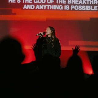 A female performer is singing into a microphone on stage. Her background is illuminated with bright red and orange lights. An audience is visible in the foreground, partially silhouetted and appearing engaged, with some hands raised. Text is displayed on a screen behind her.