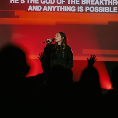 A female performer is singing into a microphone on stage. Her background is illuminated with bright red and orange lights. An audience is visible in the foreground, partially silhouetted and appearing engaged, with some hands raised. Text is displayed on a screen behind her.