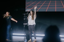 Two people are standing on a stage singing into microphones. They appear to be engaging with an audience, with one person having their arm outstretched and the other raising a hand. The backdrop features a digital display with a red grid pattern. There is stage lighting and a bottle of water is visible on the floor.