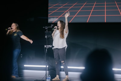 Two people are standing on a stage singing into microphones. They appear to be engaging with an audience, with one person having their arm outstretched and the other raising a hand. The backdrop features a digital display with a red grid pattern. There is stage lighting and a bottle of water is visible on the floor.