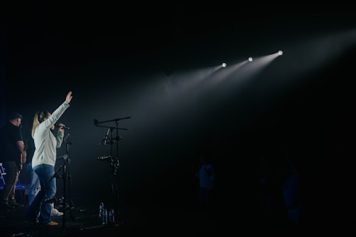 A woman is on stage holding a microphone, with her other hand raised as she sings or speaks. There are multiple microphones on stands around her. Stage lights create beams directed towards her, and other people are partially visible in the dimly lit background.