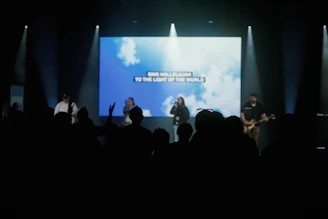 A group of people joyfully singing worship songs in a softly lit church room with pastel sky blue and white decorations.
