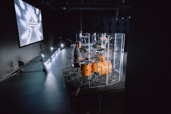 A band performing on stage with a focus on the drummer playing an orange drum set enclosed in a transparent sound shield. The stage is dimly lit with spotlights, and a large screen in the background displays text with an artistic explosion graphic. Multiple musicians are visible, including guitarists.