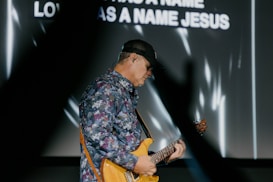 A musician wearing a floral shirt and a cap plays an electric guitar. The background features blurred text with white light patterns. The scene is dynamic and suggests a live performance.