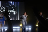 A group of people are performing with musical instruments on a stage under dim lighting. One person is singing into a microphone while another plays a guitar and a large screen in the background displays the words 'Love has a name, Love has a name Jesus.' Hands from the audience are raised in the foreground, suggesting audience participation.