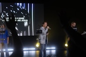 A group of people are performing with musical instruments on a stage under dim lighting. One person is singing into a microphone while another plays a guitar and a large screen in the background displays the words 'Love has a name, Love has a name Jesus.' Hands from the audience are raised in the foreground, suggesting audience participation.