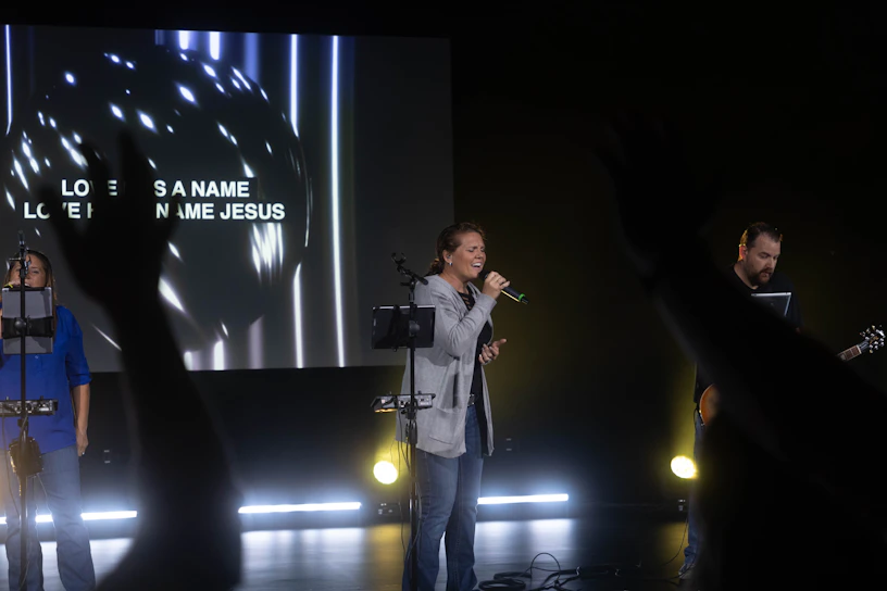 A group of people are performing with musical instruments on a stage under dim lighting. One person is singing into a microphone while another plays a guitar and a large screen in the background displays the words 'Love has a name, Love has a name Jesus.' Hands from the audience are raised in the foreground, suggesting audience participation.