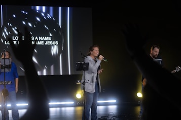 A group of people are performing with musical instruments on a stage under dim lighting. One person is singing into a microphone while another plays a guitar and a large screen in the background displays the words 'Love has a name, Love has a name Jesus.' Hands from the audience are raised in the foreground, suggesting audience participation.