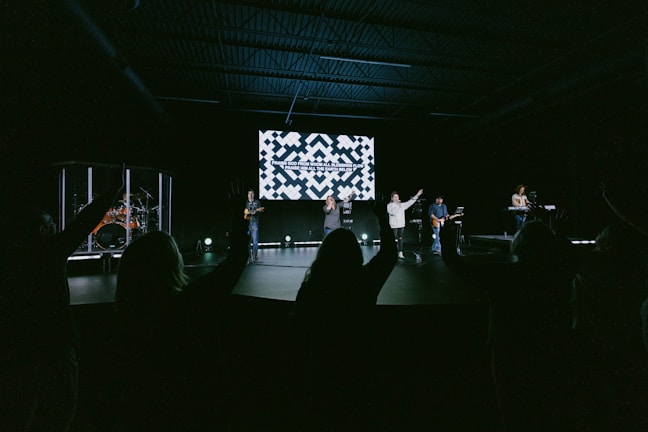 A group of musicians perform on a stage in a dimly lit auditorium. In the background, a large screen displays text. The audience is visible, silhouetted with hands raised, suggesting engagement or participation.