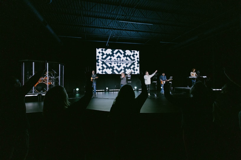 A group of musicians perform on a stage in a dimly lit auditorium. In the background, a large screen displays text. The audience is visible, silhouetted with hands raised, suggesting engagement or participation.