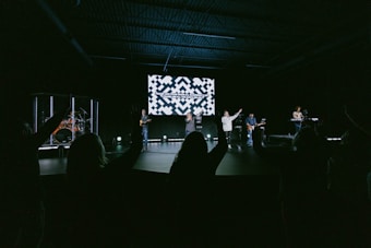 A group of musicians perform on a stage in a dimly lit auditorium. In the background, a large screen displays text. The audience is visible, silhouetted with hands raised, suggesting engagement or participation.