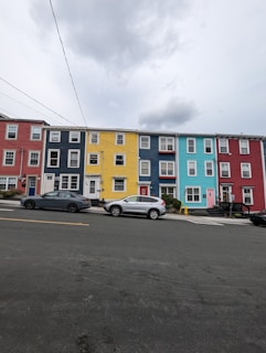 A row of colorful townhouses stands along a street. The houses are painted in vibrant shades of red, blue, yellow, and teal. A few cars are parked along the roadside in front of the houses. Overhead, utility lines stretch across the street under a cloudy sky.