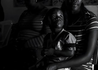 A child is sitting on an adult's lap in a dimly lit room. The adult is partially visible, with only parts of their face and upper body shown. The focus is on the child, who is gazing upwards with an intent expression. Both individuals are wearing striped clothing, and they are seated on a plastic chair.