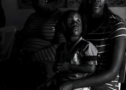 A child is sitting on an adult's lap in a dimly lit room. The adult is partially visible, with only parts of their face and upper body shown. The focus is on the child, who is gazing upwards with an intent expression. Both individuals are wearing striped clothing, and they are seated on a plastic chair.
