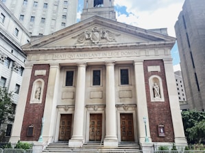 A classical building with a grand entrance characterized by large columns and intricate architectural details. The facade combines brick and stone elements, featuring Latin inscriptions and recessed statues on either side. The structure is flanked by other urban buildings and greenery is visible at the bottom left.