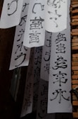 Vertical white banners with bold black calligraphy characters hang between brick walls. The banners are arranged in layers, creating a visually dynamic scene. Light and shadow play across the surfaces, emphasizing the texture of both the paper and the bricks.