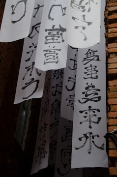 Vertical white banners with bold black calligraphy characters hang between brick walls. The banners are arranged in layers, creating a visually dynamic scene. Light and shadow play across the surfaces, emphasizing the texture of both the paper and the bricks.