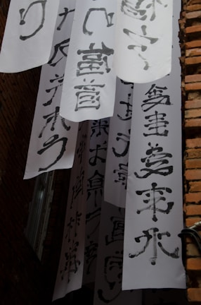 Vertical white banners with bold black calligraphy characters hang between brick walls. The banners are arranged in layers, creating a visually dynamic scene. Light and shadow play across the surfaces, emphasizing the texture of both the paper and the bricks.