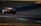 A sports car with GT3 markings is racing on a track with visible tire marks. The car is painted in red, black, and white, and features a prominent rear wing. The track is surrounded by grass, and the lighting suggests it is late afternoon or early evening.
