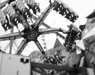 A black and white photo of an amusement park ride in motion, featuring spinning arms and seats with passengers. The ride is composed of multi-segmented arms attached to a central hub, with seats aligned in various directions. The background includes blurred structures and possibly other rides, conveying a sense of speed and excitement.