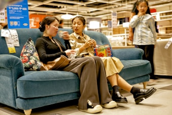 Two women are sitting on a blue sofa in a store, engaged in conversation. One is holding a snack and gesturing with her hand, while the other listens attentively. A box with colorful graphics is placed between them. A young girl stands nearby, looking on. Price tags are visible on the sofa, suggesting a shopping setting.