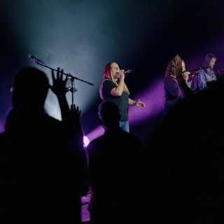 A group of performers are singing on stage with microphones. The stage is illuminated by purple and blue lights, creating a dramatic atmosphere. Several people are visible in the foreground, silhouetted against the bright stage light, with some raising their hands.
