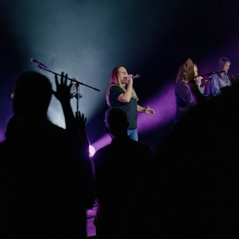 A group of performers are singing on stage with microphones. The stage is illuminated by purple and blue lights, creating a dramatic atmosphere. Several people are visible in the foreground, silhouetted against the bright stage light, with some raising their hands.