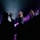 A group of performers are singing on stage with microphones. The stage is illuminated by purple and blue lights, creating a dramatic atmosphere. Several people are visible in the foreground, silhouetted against the bright stage light, with some raising their hands.