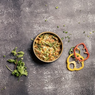 A bowl of richly seasoned curry garnished with fresh cilantro and thinly sliced green vegetables is placed on a textured, dark surface. Next to the bowl are slices of red, yellow, and green bell peppers, along with sprigs of fresh mint and cilantro scattered nearby.