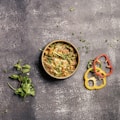 A bowl of richly seasoned curry garnished with fresh cilantro and thinly sliced green vegetables is placed on a textured, dark surface. Next to the bowl are slices of red, yellow, and green bell peppers, along with sprigs of fresh mint and cilantro scattered nearby.