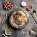 A plate of flavorful biryani garnished with roasted meat, a whole garlic bulb, and a charred tomato sits on a rustic background. Surrounding the main dish are small bowls containing condiments like yogurt, red sauce, spicy green soup, and gulab jamun. A sprig of mint and lime slices are arranged nearby, with cutlery placed neatly to the side.