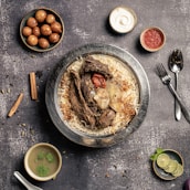 A plate of flavorful biryani garnished with roasted meat, a whole garlic bulb, and a charred tomato sits on a rustic background. Surrounding the main dish are small bowls containing condiments like yogurt, red sauce, spicy green soup, and gulab jamun. A sprig of mint and lime slices are arranged nearby, with cutlery placed neatly to the side.