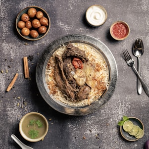 A plate of flavorful biryani garnished with roasted meat, a whole garlic bulb, and a charred tomato sits on a rustic background. Surrounding the main dish are small bowls containing condiments like yogurt, red sauce, spicy green soup, and gulab jamun. A sprig of mint and lime slices are arranged nearby, with cutlery placed neatly to the side.