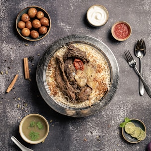A plate of flavorful biryani garnished with roasted meat, a whole garlic bulb, and a charred tomato sits on a rustic background. Surrounding the main dish are small bowls containing condiments like yogurt, red sauce, spicy green soup, and gulab jamun. A sprig of mint and lime slices are arranged nearby, with cutlery placed neatly to the side.