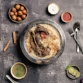 A plate of flavorful biryani garnished with roasted meat, a whole garlic bulb, and a charred tomato sits on a rustic background. Surrounding the main dish are small bowls containing condiments like yogurt, red sauce, spicy green soup, and gulab jamun. A sprig of mint and lime slices are arranged nearby, with cutlery placed neatly to the side.